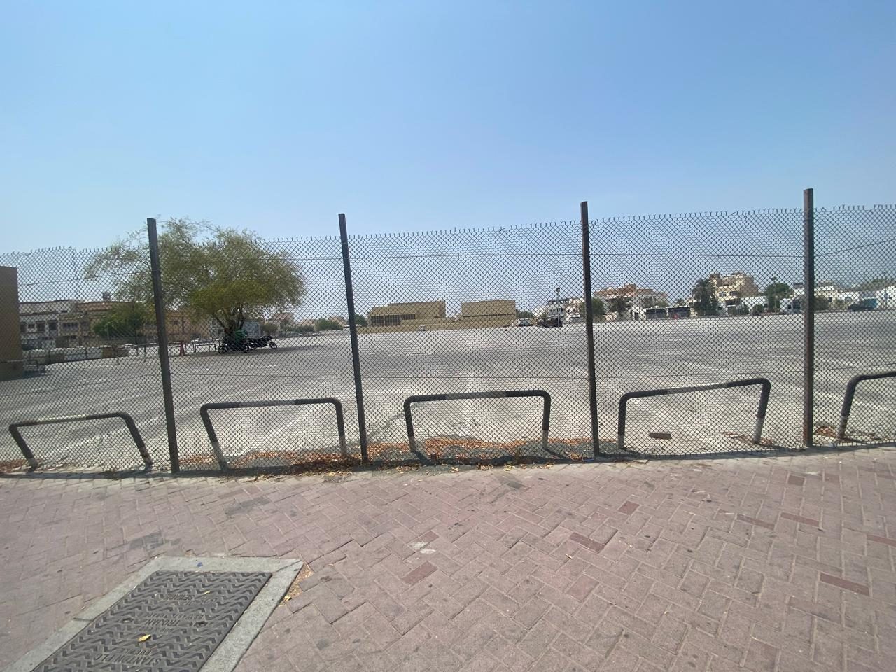 The cleared, level plot seen from behind the boundary fence, with low-rise buildings beyond